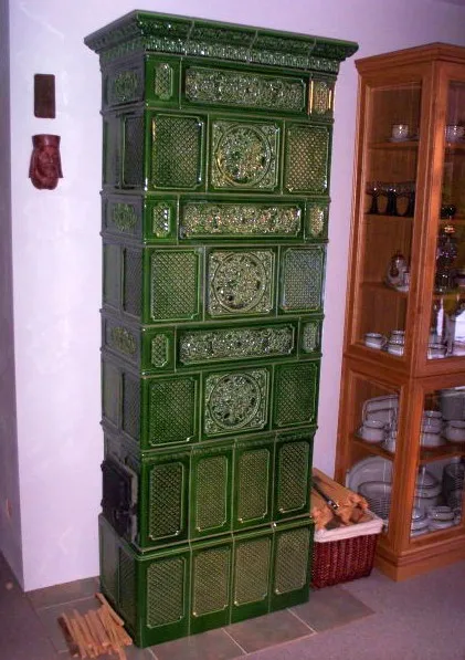 Vysoká tmavě zelená kachlová kamna s&nbsp;reliéfními kachlemi vedle prosklené vitríny