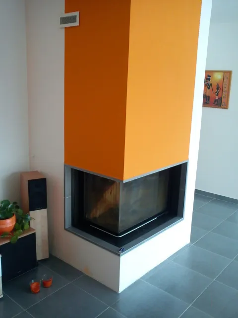 Moderní rohový krb s&nbsp;prosklením do dvou stran a&nbsp;oranžově omítnutým tělesem