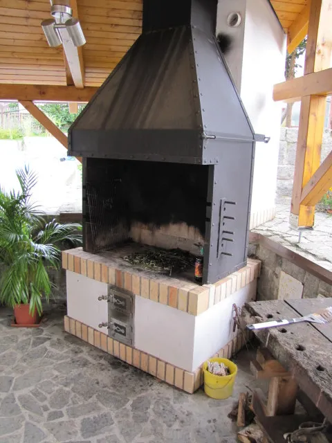 Zděný venkovní gril s&nbsp;kovovým odtahem pod dřevěnou pergolou